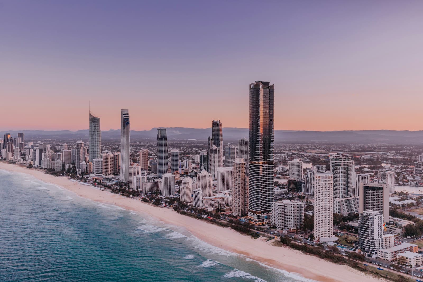 Aerial-style or elevated view of Burleigh Heads headland and beach at sunrise, pandanus trees in foreground, long crescent of sand stretching north toward Surfers, no people close to camera. Not a Surfers Paradise night-skyline shot.