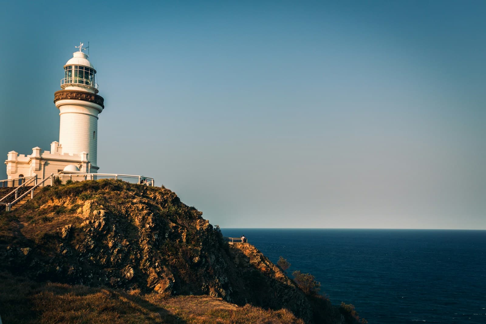 Small group at Cape Byron lookout just before sunrise. Lighthouse silhouetted, ocean still slate, sky turning peach. Arakwal guide gesturing toward the headland. Quiet, contemplative.