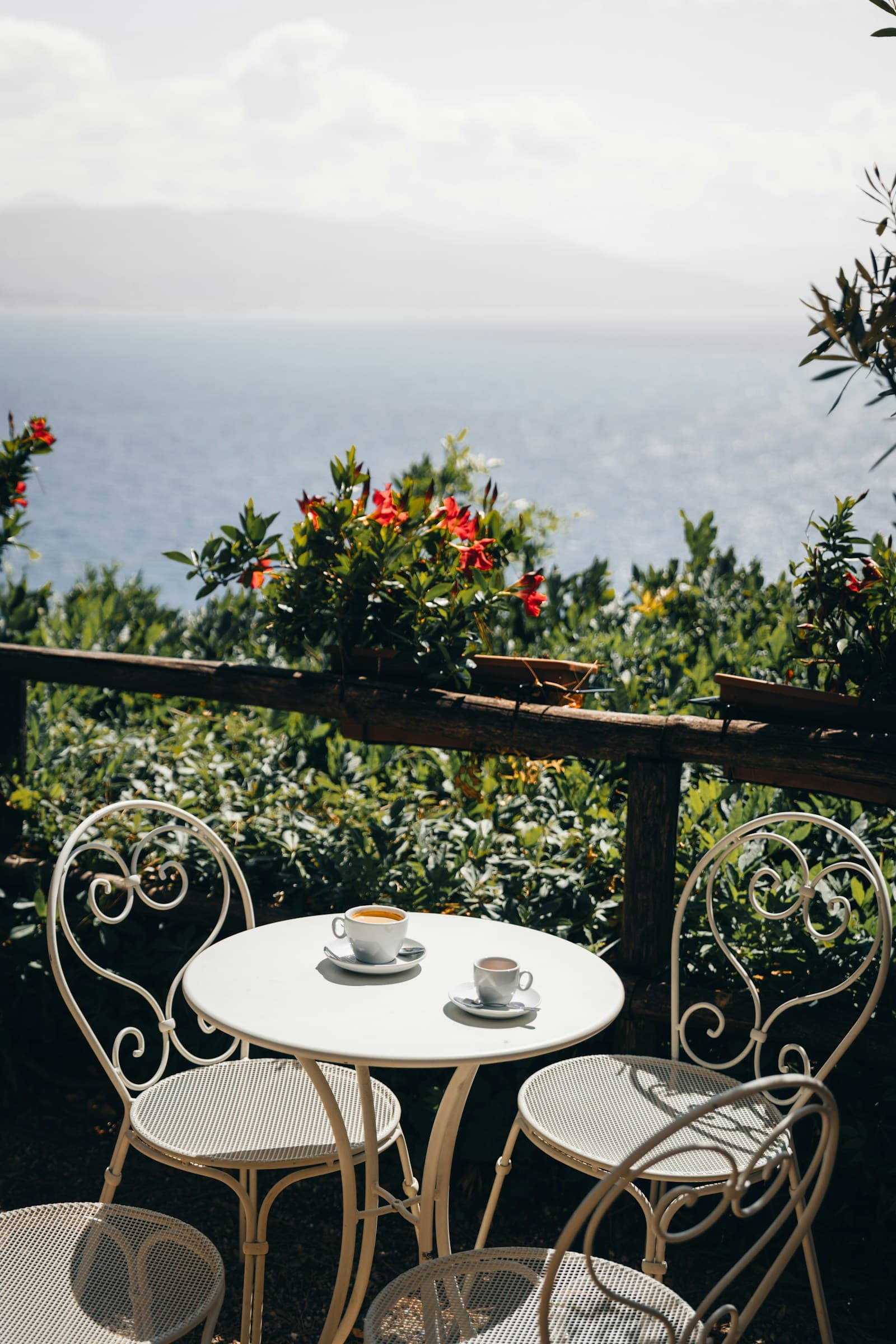 A small, unfussy cafe terrace in Sunshine Beach in late-morning light, two locals at a timber table mid-conversation over coffee, bare feet, boardshorts, no branding visible. Shot slightly from above with a mid-lens, warm.